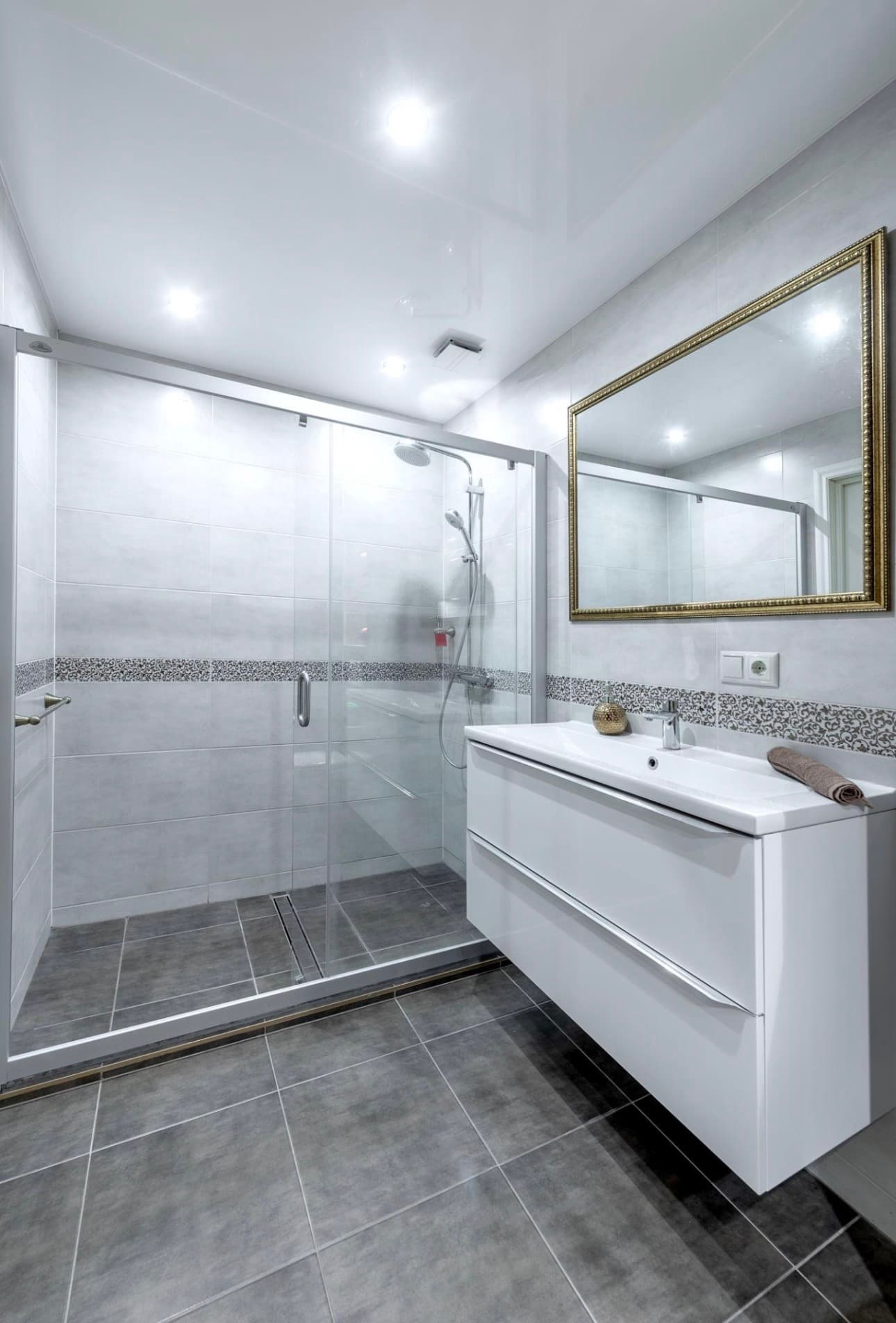 Clean bathroom with gray tile floor, glass sliding shower with mosaic accent strip, and gold-framed mirror